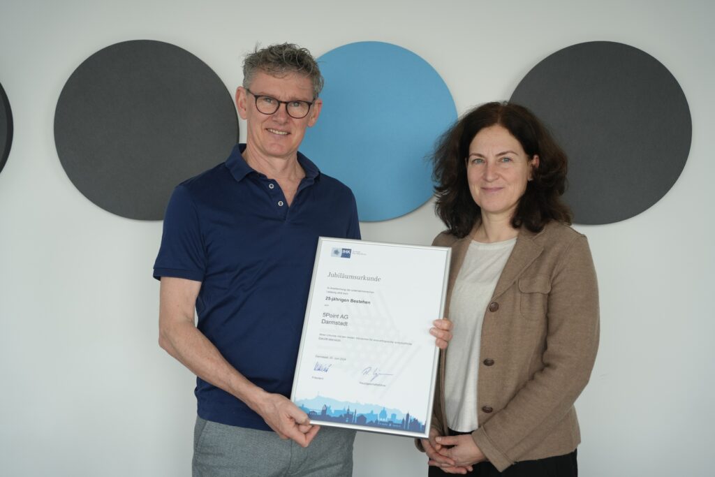 Thorsten Lenk und eine Frau von der IHK die eine Urkunde zum 25-jährigen Bestehen der 5 Point AG überreicht hat vor einer weißen Wand mit dem Logo der 5 Point AG im Hintergrund